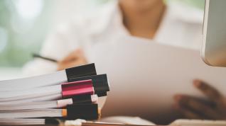 Binder clipped stacks of papers in front of a person holding a piece of paper near a computer screen.