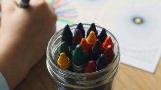 Crayons in a mason jar, a child’s arm is holding a crayon and they are drawing on a piece of paper. 